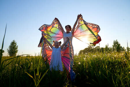 Mother and daughter with bright fabric like butterfly wings play, fly, run, jump in meadow or field with green grass and dandelion flowers. Young woman and a little girl have fun on natural landscapeの写真素材