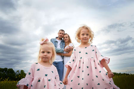 A family including two young parents and daughters on a walk in a meadow with grass and flowers. Dad, mom, girls relaxing and having fun in nature on a summer day with cloudsの写真素材