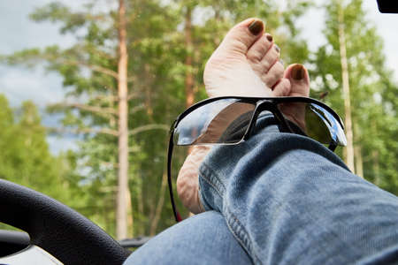 View on leg of woman with black sunglasses lying on the dashboard of a car near the steering wheel and the forest in the background behind the windshield. The concept of travel and leisure in natureの写真素材