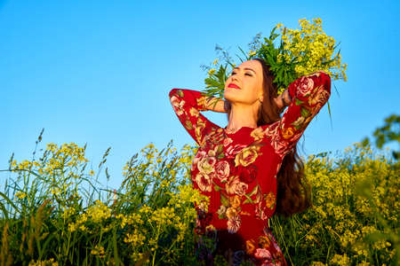 Beautiful woman with long hair in red dress holding wreath of yellow flowers and enjoying summer...