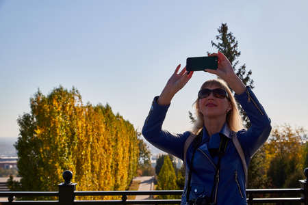 Attractive beautiful young girl holding cell phone in autumn park and making selfie. ... Female...