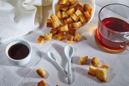 Square toasted pieces of homemade delicious rusk, hardtack, Dryasdust, zwieback, Liquid honey in a saucer and black tee in a cap on a white tableclothの写真素材