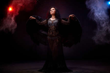 Girl in black dress and with black wings posing in dark studo during photoshoot with flour or dust and light. Dangerous witch during global struggle between good and evilの写真素材