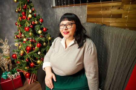 Fat cute woman with glasses and black hair posing in a room decorated for Christmas or New year in...