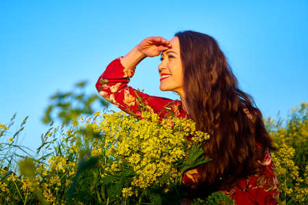 Beautiful woman with long hair in red dress holding bouquet of yellow flowers and enjoying summer sunny day in green field with green grassの写真素材