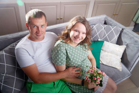 Cute couple including a pregnant woman and a caring man in the living room together. Beautiful pregnant future mother and father at homeの写真素材