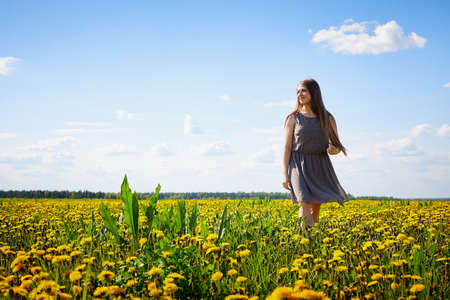 Beautiful young woman on a field with green grass and yellow dandelion flowers in a sunny day. Girl on nature with yellow flowers and blue skyの写真素材