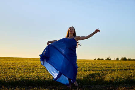 Beautiful woman or girl with magnificent figure and plastic movements walking and dancing in green field with trimmed grass in the setting sun during sunset with warm yellow lightの写真素材
