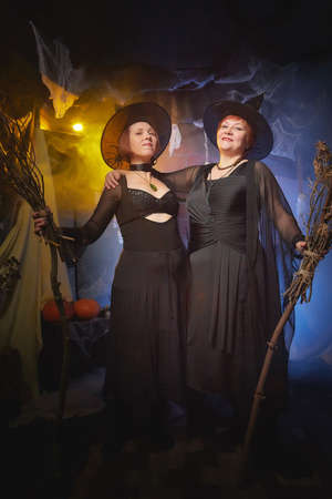 Two adult women looking as witches having fun and posing in dark room decorated for Halloween during...