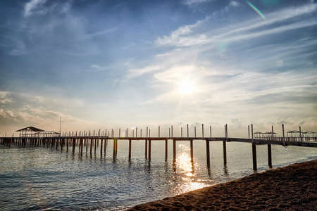 View from sand beach to water of sea, waves and pier in a nice day or evening with blues sky, bright sun and white clouds. The concept of a holiday on the sea or ocean in the Southの写真素材