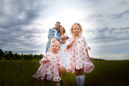 Family including two young parents and daughters on a walk in a meadow with grass and flowers. Dad, mom, girls relaxing and having fun in nature on a summer day with cloudsの写真素材