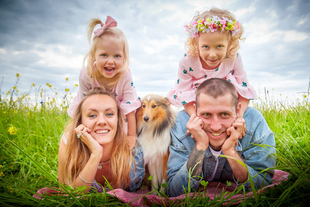 Family including two young parents and daughters on a walk in a meadow with grass and flowers. Dad, mom, girls relaxing and having fun in nature on a summer day with cloudsの写真素材