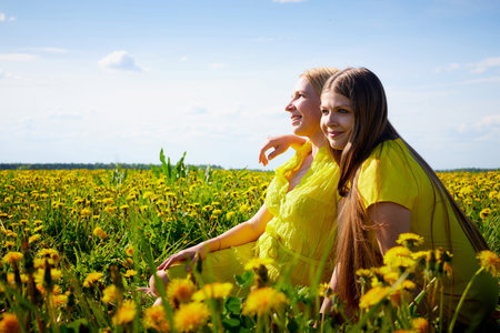 Two pretty women on a field with green grass and yellow dandelion flowers in a sunny day with blue sky. Girls having rest and fun on nature. Female friends or sisters resting togetherの写真素材