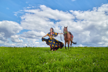 Beautiful gypsy girl walking with a horse in a field with green glass in summer day and blue sky and white clouds background. Model in ethnic dress posing with farm animalの写真素材