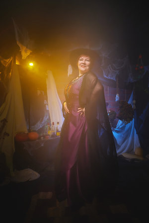 Slim witch in maroon dress and black hat in room decorated for Halloween