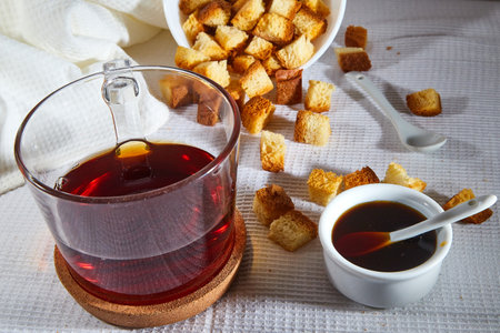 Square toasted pieces of homemade delicious rusk, hardtack, Dryasdust, zwieback, Liquid honey in a saucer and black tee in a cap on a white tableclothの写真素材