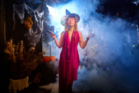 Blonde witch in red dress and black hat in Halloween decoration indoors with cell phone and smoke near her. Woman taking selfie during holiday carnivalの写真素材