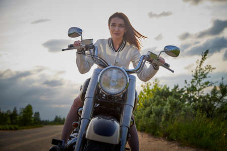 Attractive brunette motorcyclist with motorcycle in a summer evening during sunset. Adventure and...