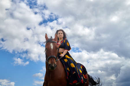 Beautiful gypsy girl on a horse in a field with green glass in summer day and blue sky and white...