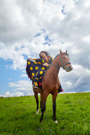 Beautiful gypsy girl on a horse in a field with green glass in summer day and blue sky and white...