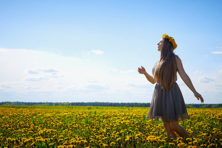 Beautiful young woman on a field with green grass and yellow dandelion flowers in a sunny day. Girl on nature with yellow flowers and blue skyの写真素材
