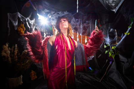 Blonde witch in red dress and black hat in Halloween decoration indoors with cell phone. Woman taking selfie during holiday carnivalの素材 [FY310164645554]