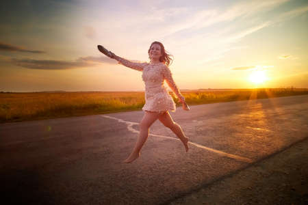 Beautiful sexy girl in pink dress and with high-heeled shoes in hand walking on the asphalt highway during sunset and field with grass on side of the road.の写真素材