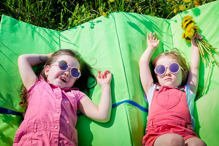 Two small girls on a green rubber mattress in a field with yellow dandelions. Sisters having rest in spring in natureの写真素材