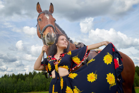 Portrait of beautiful gypsy girl with a horse on a field with green glass in summer day and blue sky...