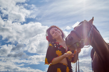 Portrait of beautiful gypsy girl with a horse on a field with green glass in summer day and blue sky...