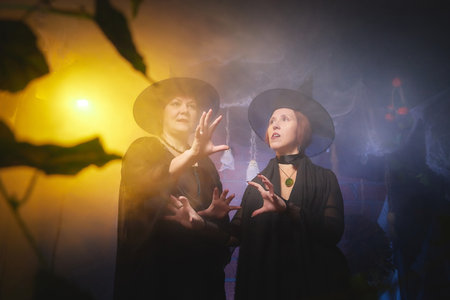 Two adult women looking as witches having fun and posing in dark room decorated for Halloween during photoshoot in studioの写真素材
