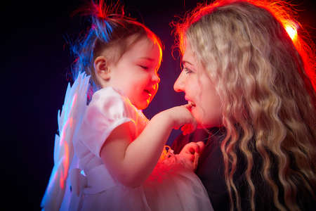 A beautiful blonde mother with curly hair and cute small girl with white wings looking like a nice angels in photo shoot in studio with black background and color lightの写真素材