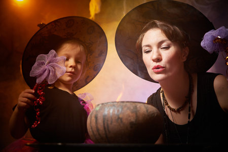 Beautiful brunette mother and cute little daughter looking as witches in special dresses and hats conjuring with a pot in room decorated for Halloween. Halloween style photo shootの写真素材