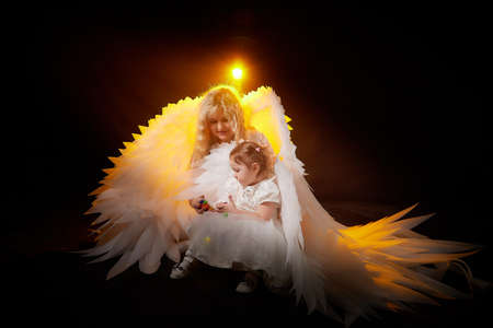 A beautiful blonde mother with curly hair and cute small girl with white wings looking like nice angels in photoshoot in studio with black backgroundの写真素材