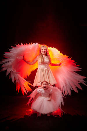 A beautiful blonde mother with curly hair and cute small girl with white wings looking like nice angels in photoshoot in studio with black backgroundの写真素材
