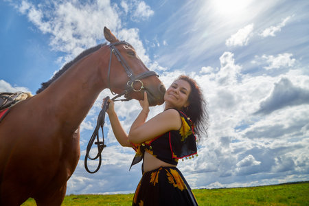 Portrait of beautiful gypsy girl with a horse on a field with green glass in summer day and blue sky...