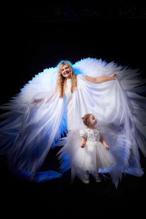 A beautiful blonde mother with curly hair and cute small girl with white wings looking like nice angels in photoshoot in studio with black backgroundの写真素材