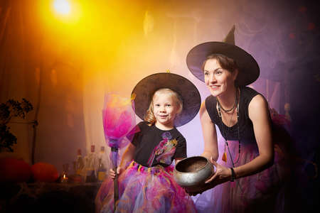 Beautiful brunette mother and cute little daughter looking as witches in special dresses and hats...