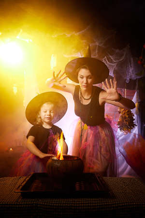 Beautiful brunette mother and cute little daughter looking as witches in special dresses and hats conjuring with a pot in room decorated for Halloween. Halloween style photo shootの写真素材
