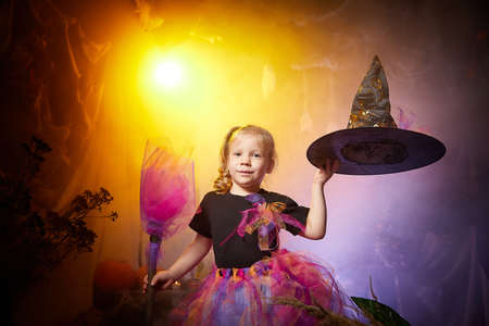 Little cute blonde girl looking as witch in special dress and hat in room decorated for Halloween. Witchcraft and wizardry in carnival. Halloween style photo shootの写真素材
