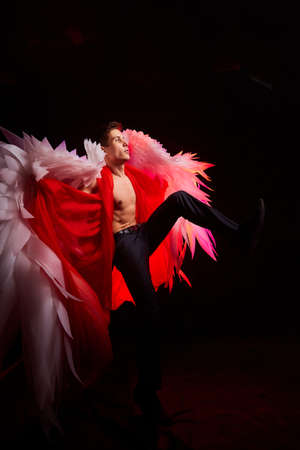 Handsome young athletic man with a bare torso who looks like an angel with white wings. Model dancer posing in dark studio on black backgroundの写真素材