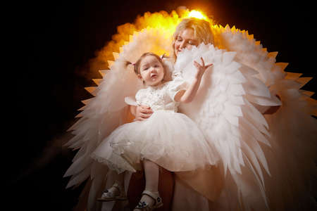 A beautiful blonde mother with curly hair and cute small girl with white wings looking like nice angels in photoshoot in studio with black backgroundの写真素材