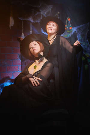 Two adult women looking as witches having fun and posing in dark room decorated for Halloween during photoshoot in studio.の写真素材