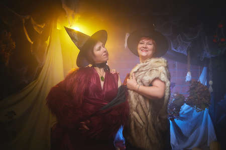Two adult women looking as witches having fun and posing in dark room decorated for Halloween during photoshoot in studio.の写真素材