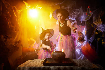 Beautiful brunette mother and cute little daughter looking as witches in special dresses and hats conjuring with a pot in room decorated for Halloween. Halloween style photo shoot.の写真素材