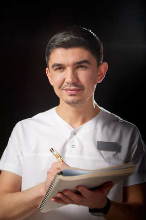 A young man who is a medical doctor in a white uniform poses against a black background in the studio with a notepad in handの写真素材