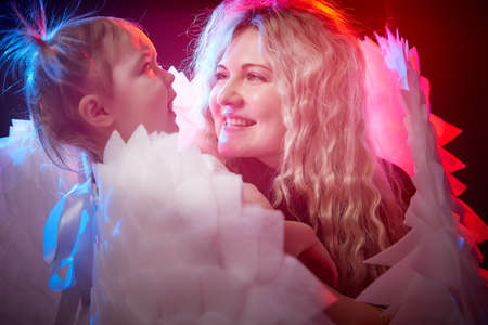 A beautiful blonde mother with curly hair and cute small girl with white wings looking like a nice angels in photo shoot in studio with black background and color lightの写真素材