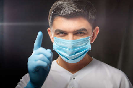 A young man who is a medical doctor in a medical mask a white uniform poses against a black background in the studio.の写真素材