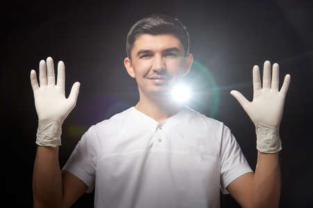 A young man who is a medical doctor in a white medical gloves and in uniform poses against a black background in the studio. Medic during the OKVED-19 pandemicの写真素材