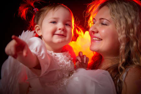 A beautiful blonde mother with curly hair and cute small girl with white wings looking like a nice angels in photo shoot in studio with black background and color lightの写真素材